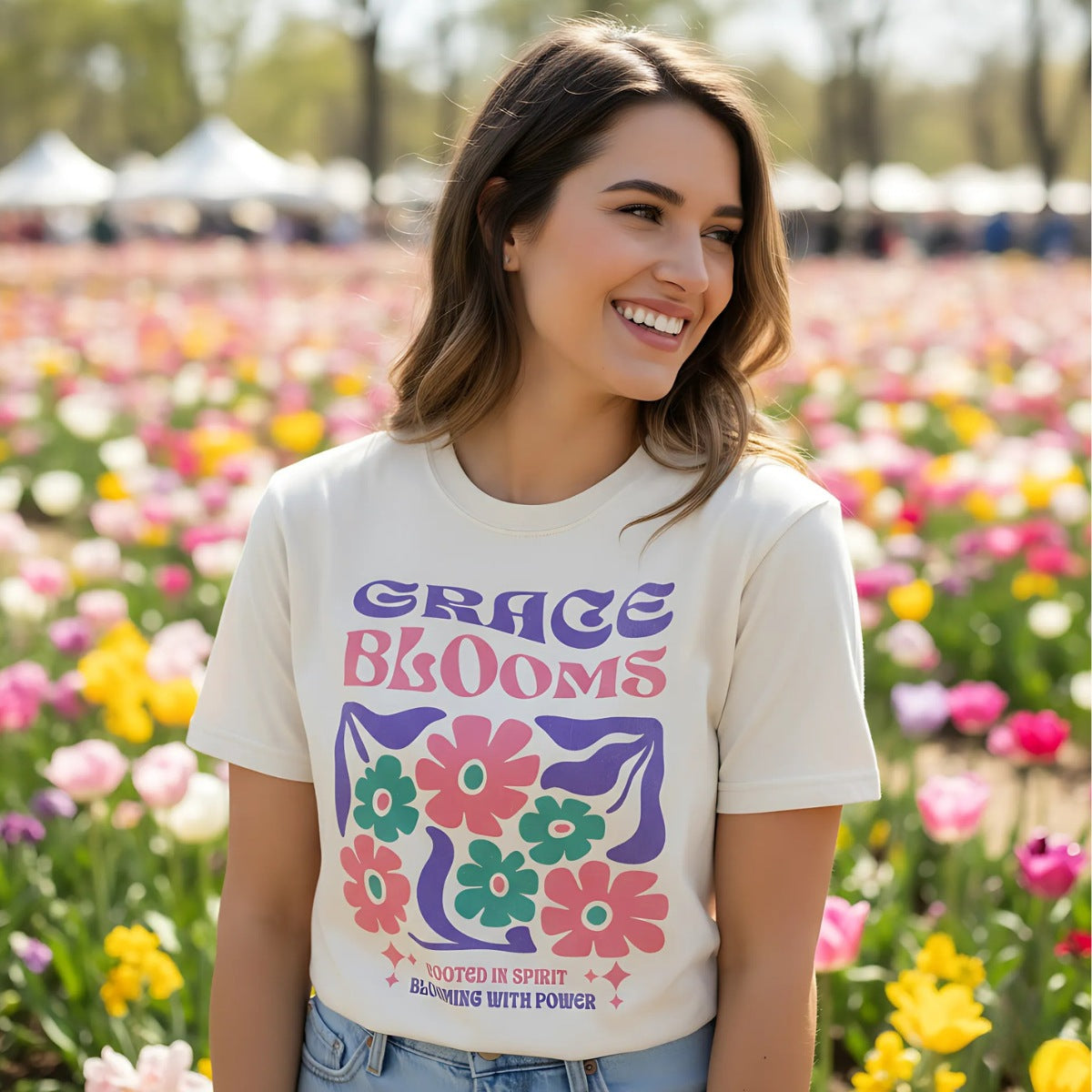 "Young woman smiling in a field of tulips, wearing ivory Comfort Colors 1717 Christian T-shirt with bold retro floral design and text ‘Grace Blooms — Rooted in Spirit, Blooming with Power.’ Vibrant Christian apparel lifestyle photography."
