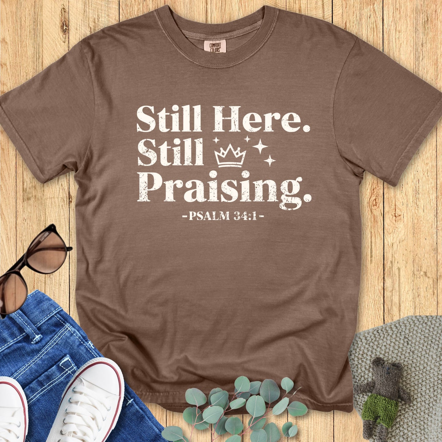 Comfort Colors 1717 Espresso brown Christian T-shirt with the text ‘Still Here. Still Praising. -Psalm 34:1’ in white distressed font, styled with jeans, sunglasses, and sneakers on a wooden background