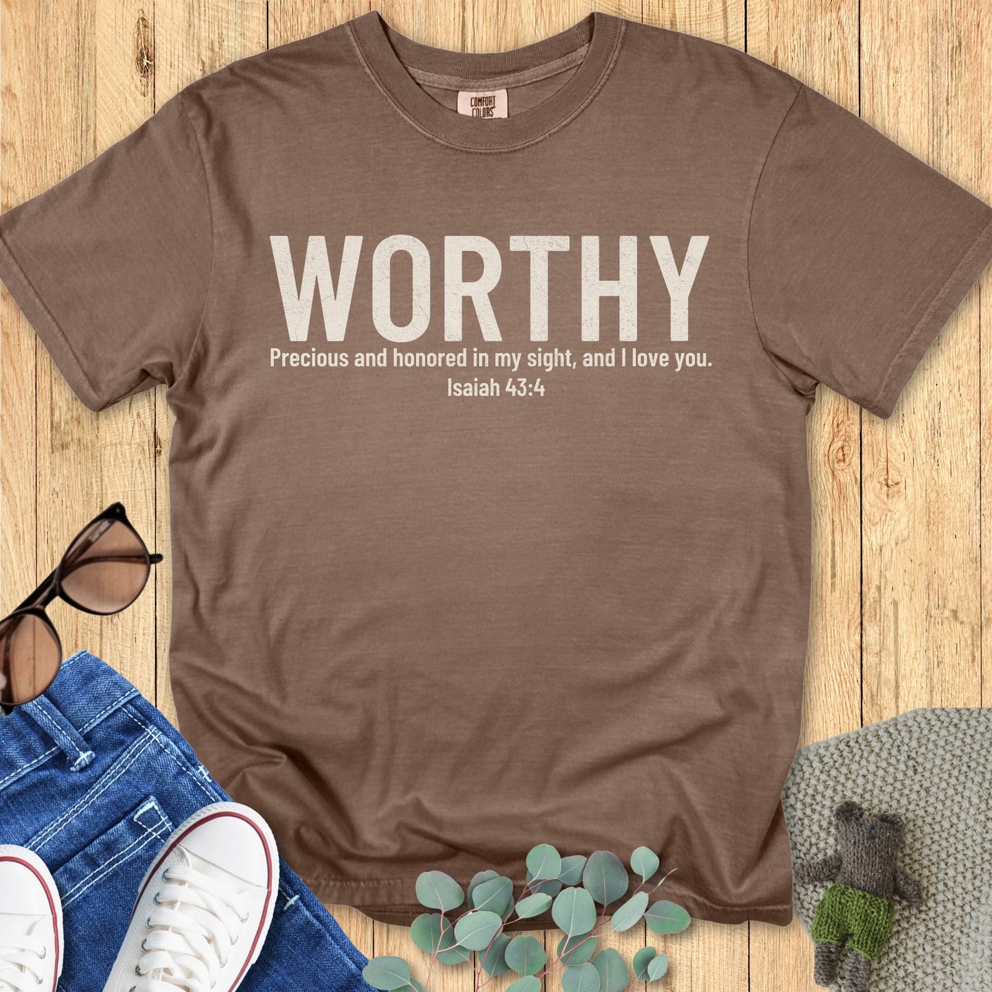 Comfort Colors 1717 espresso brown Christian women’s t-shirt with the word “WORTHY” in bold white text. Below, the verse Isaiah 43:4 reads: “Precious and honored in my sight, and I love you.” Laid flat with jeans, sunglasses, and greenery on wood background
