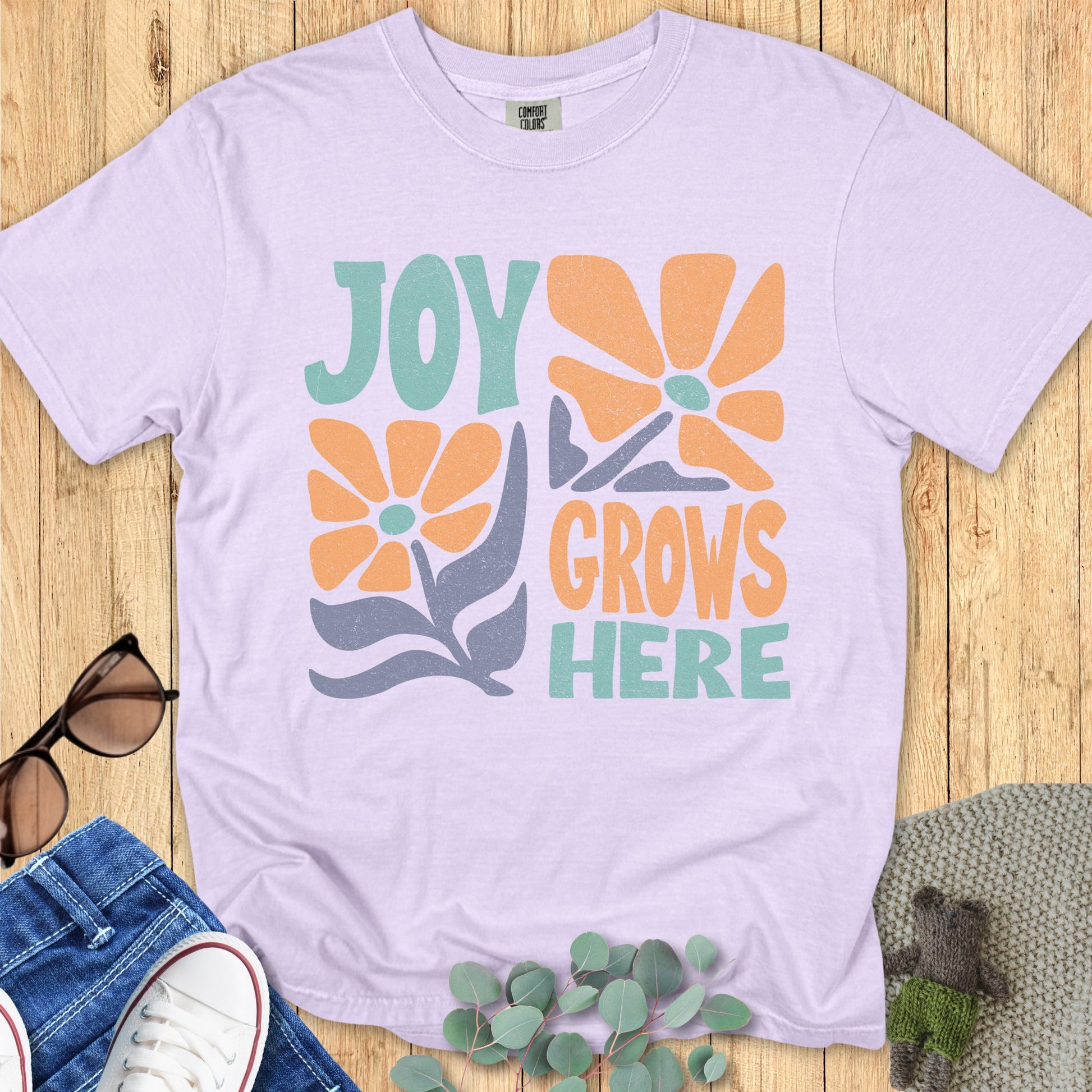 Flat lay of a light purple T-shirt featuring the “Joy Grows Here” text and flower design, arranged with casual accessories on a wooden backdrop