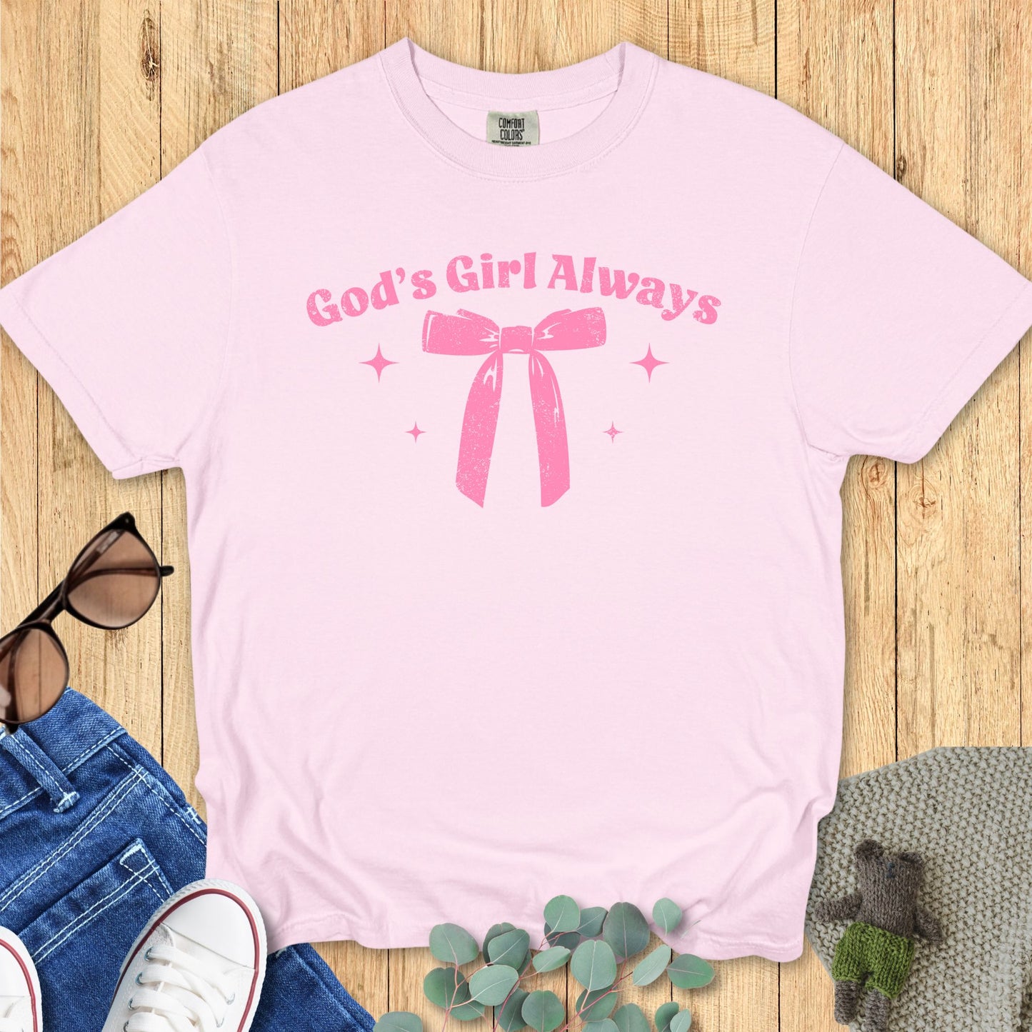 lat lay of a light pink T-shirt featuring the same “God’s Girl Always” pink bow and sparkles design.