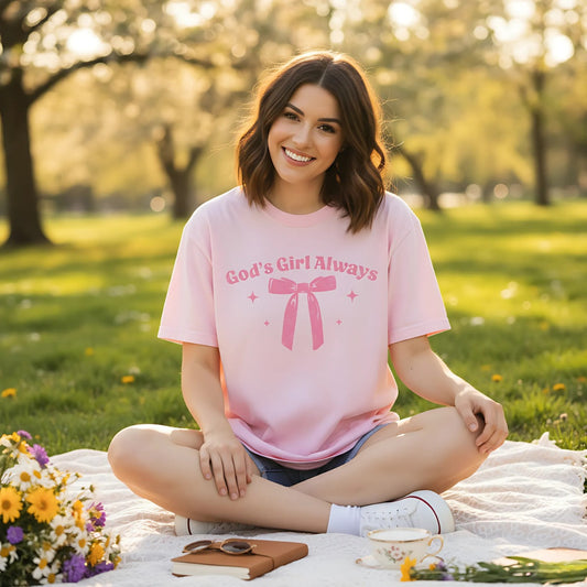 Woman sitting on a picnic blanket outdoors, smiling and wearing a light pink T-shirt with the phrase “God’s Girl Always” above a pink bow and sparkle