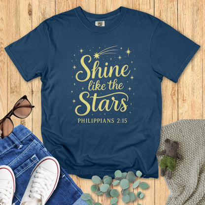 Flat lay of a midnight blue Comfort Colors 1717 T-shirt with the Christian design “Shine like the Stars – Philippians 2:15” in golden yellow, paired with jeans and casual accessories on a wooden floor.