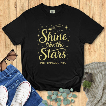 Flat lay of a black Comfort Colors 1717 T-shirt with the design “Shine like the Stars – Philippians 2:15” in gold text surrounded by stars and a comet, styled with jeans, white sneakers, and sunglasses on a wooden background.