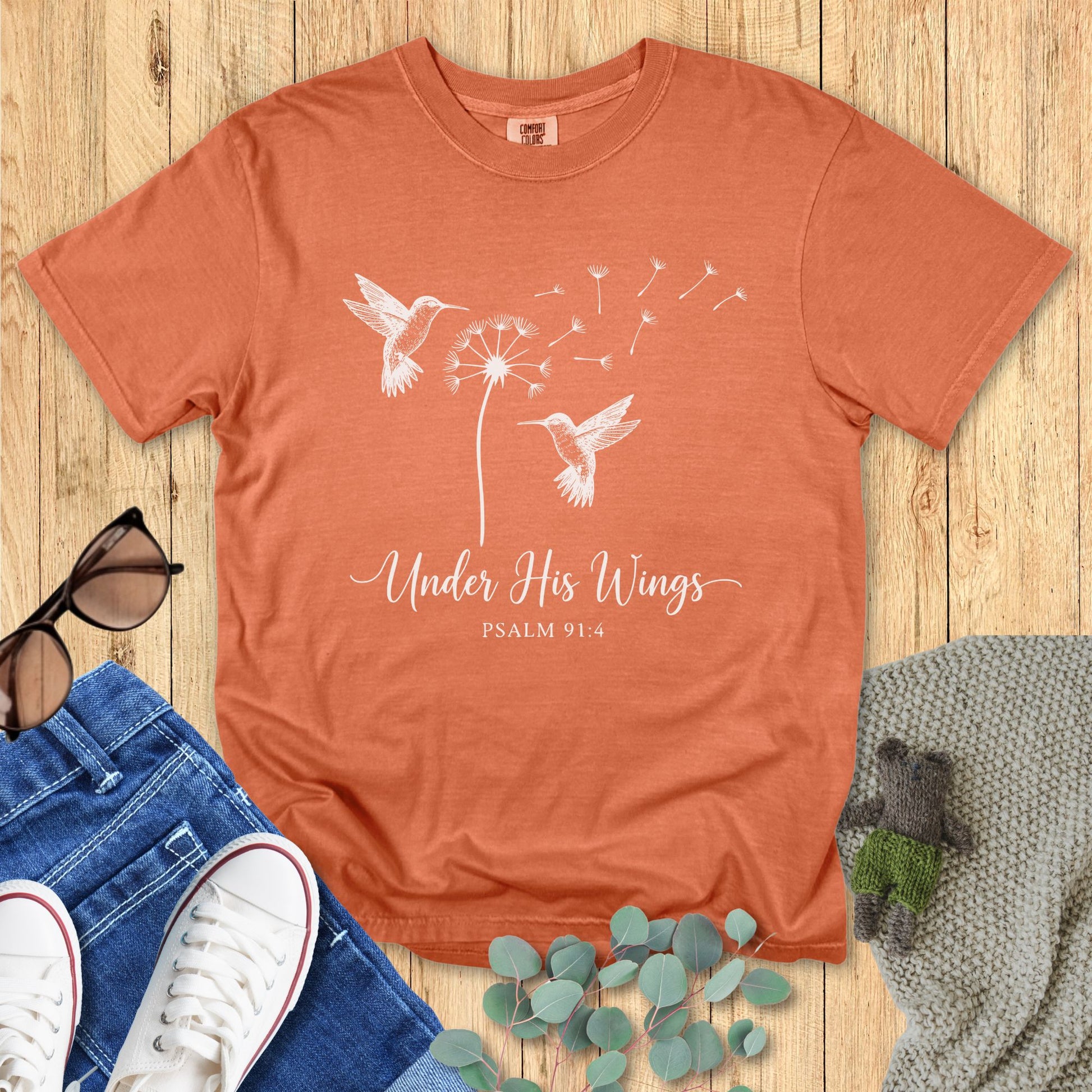 Yam Comfort Colors Christian T-shirt with white hummingbird and dandelion artwork, and verse "Under His Wings Psalm 91:4." Inspirational faith-based tee flat lay with jeans, sunglasses, and sneakers on wood floor background.