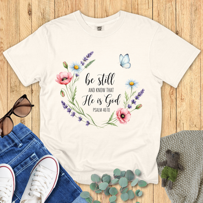 Ivory Comfort Colors Christian T-shirt with floral wreath and butterfly design featuring scripture "Be Still and Know That He Is God, Psalm 46:10." Inspirational Christian tee flat lay with jeans, sneakers, and sunglasses on wood background.