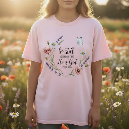 Blossom pink Comfort Colors Christian T-shirt with Bible verse "Be Still and Know That He Is God, Psalm 46:10." Floral wreath design with lavender, daisies, poppies, and a butterfly, modeled outdoors in a flower field.