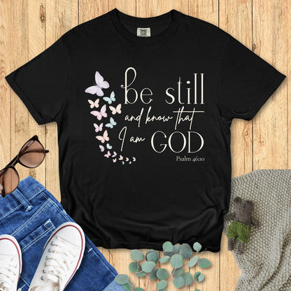 Black Comfort Colors Christian T-shirt with butterfly design and scripture "Be Still and Know That I Am God, Psalm 46:10." Faith-based inspirational tee flat lay with jeans, sneakers, and sunglasses on wood background.