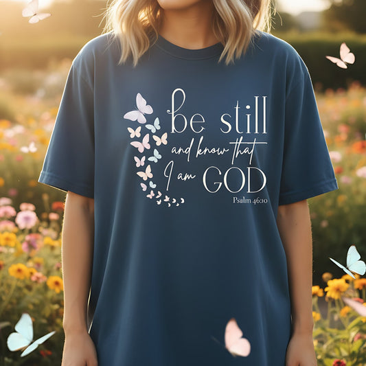 Midnight navy Comfort Colors Christian T-shirt with pastel butterfly design and verse "Be Still and Know That I Am God, Psalm 46:10." Modeled outdoors in a flower garden with butterflies flying around for a natural lifestyle look.