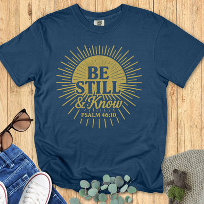 Midnight navy Comfort Colors Christian T-shirt featuring bold sunburst artwork and verse "Be Still & Know, Psalm 46:10." Faith graphic tee flat lay styled with denim, sneakers, and greenery on wood surface.