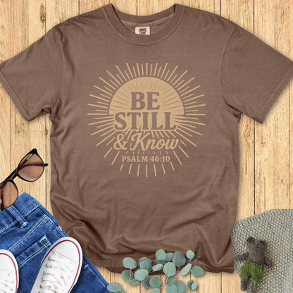 Espresso brown Comfort Colors Christian T-shirt with vintage sunburst design and scripture "Be Still & Know, Psalm 46:10." Inspirational faith-based tee flat lay with jeans, sneakers, and sunglasses on wooden background.