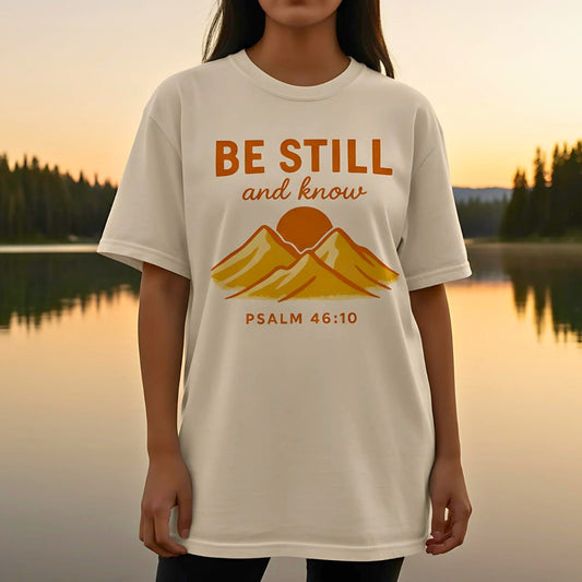 Ivory Comfort Colors Christian T-shirt with retro mountain and sun design and scripture "Be Still and Know, Psalm 46:10." Modeled outdoors at sunset near a lake for a natural lifestyle look.