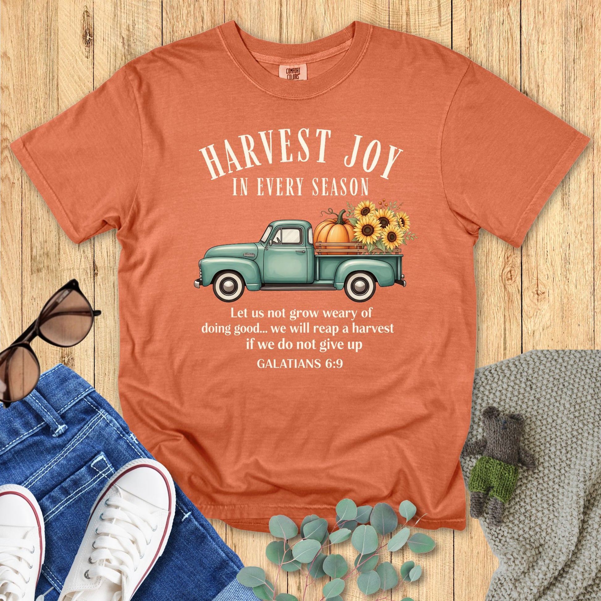Yam orange Comfort Colors Christian T-shirt with teal farm truck, pumpkins, and sunflower harvest design. Verse "Harvest Joy in Every Season – Galatians 6:9." Autumn Christian tee flat lay styled with denim and accessories.