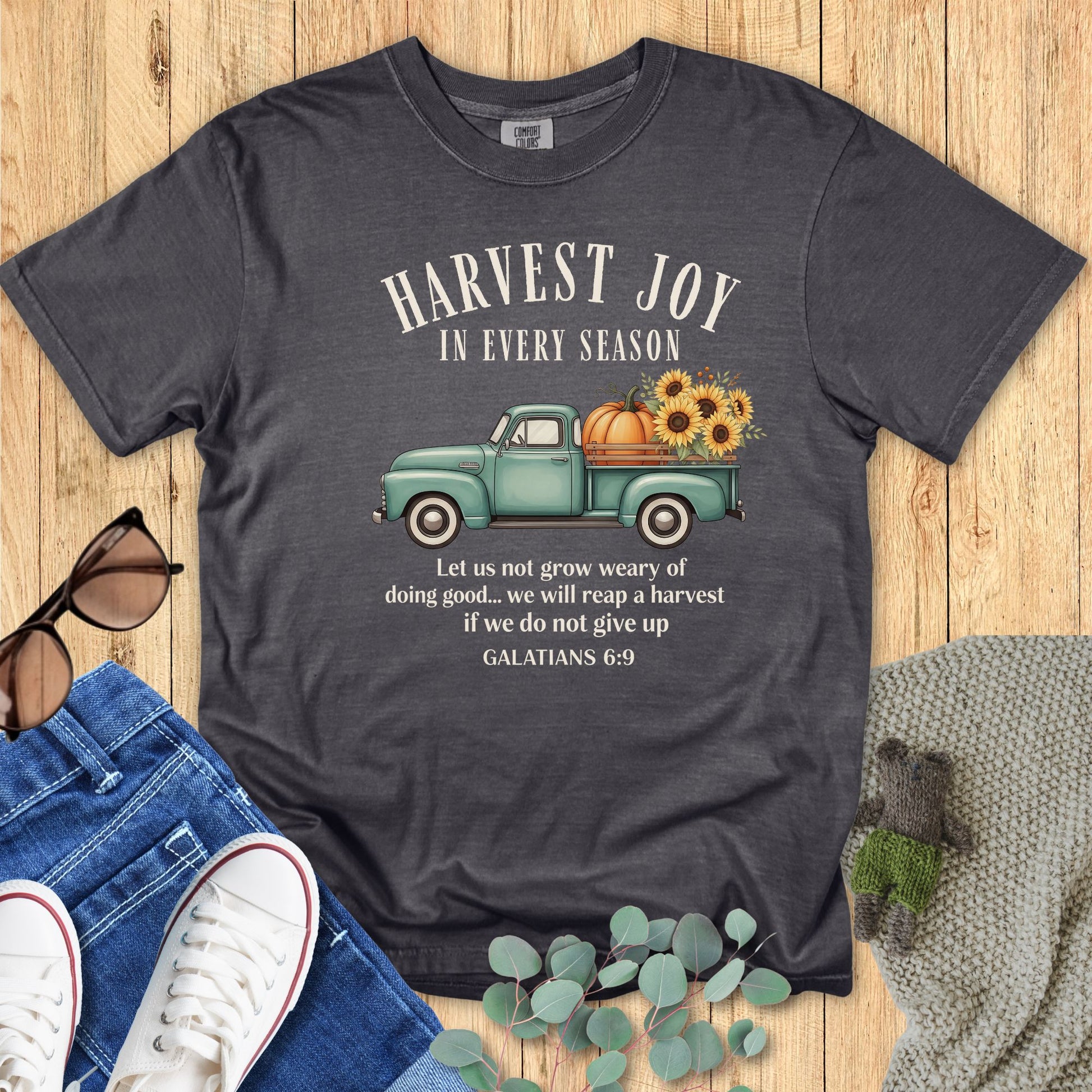 Pepper gray Comfort Colors Christian T-shirt with vintage teal truck carrying pumpkins and sunflowers, featuring scripture "Harvest Joy in Every Season – Galatians 6:9." Faith-based fall graphic tee flat lay with jeans, sneakers, and sunglasses on wooden background.