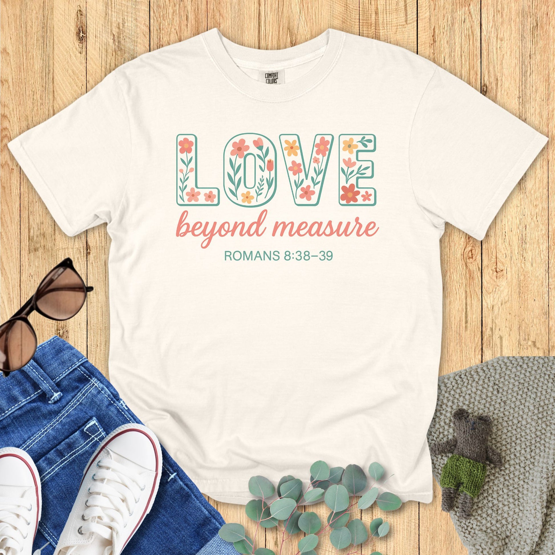Ivory Comfort Colors Christian T-shirt with floral block letters spelling "LOVE," script text "beyond measure," and Bible verse Romans 8:38–39. Faith-based inspirational tee flat lay with jeans, sneakers, and sunglasses on wood background.
