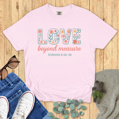 Blossom pink Comfort Colors Christian T-shirt with floral block text "LOVE beyond measure," and verse Romans 8:38–39. Faith-based inspirational tee flat lay with jeans, sneakers, and greenery on wood surface.