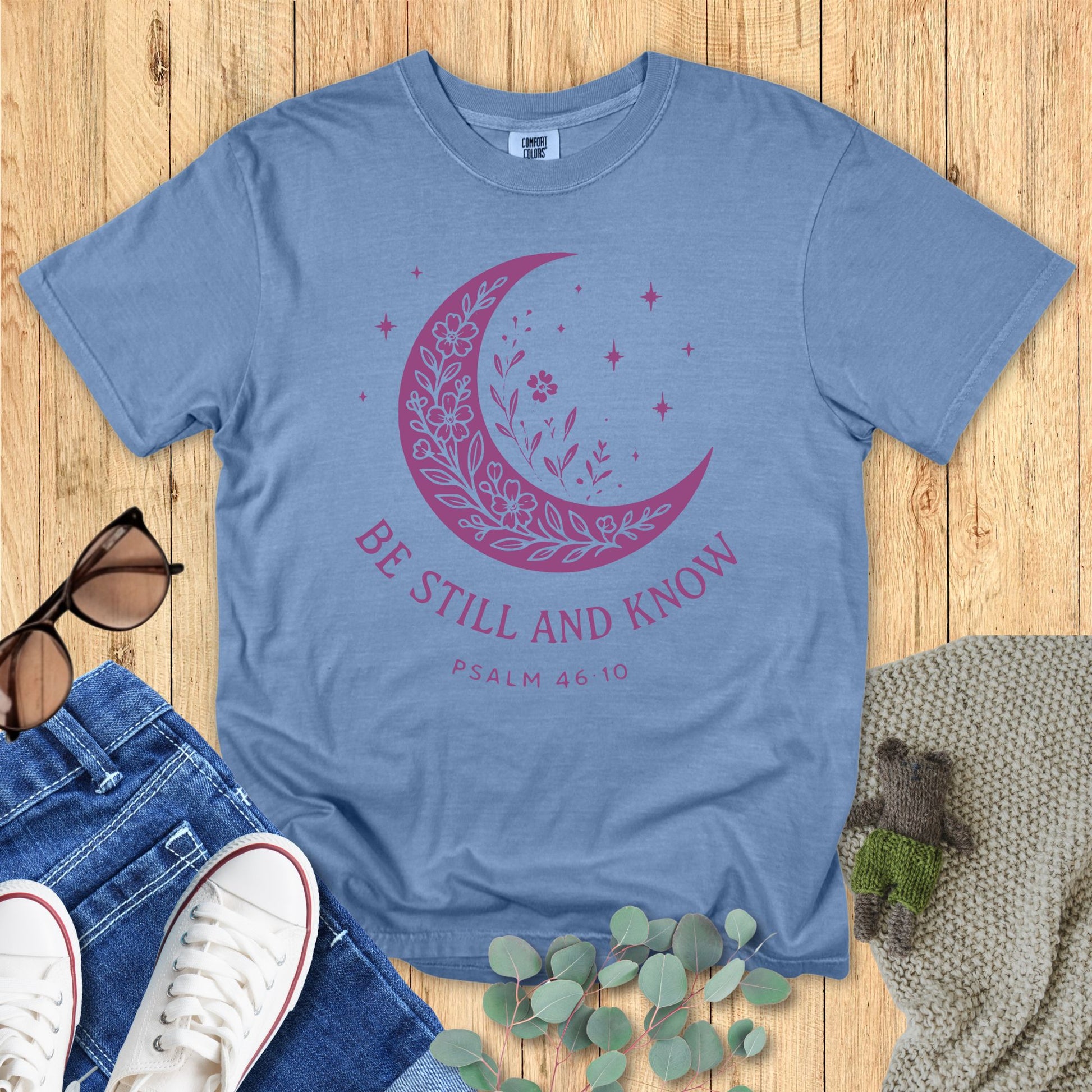 Flat lay of a Comfort Colors Christian T-shirt in blue jean, featuring a purple crescent moon filled with floral details and stars, with text “Be Still and Know – Psalm 46:10.”