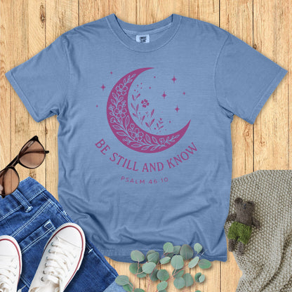 Flat lay of a Comfort Colors Christian T-shirt in blue jean, featuring a purple crescent moon filled with floral details and stars, with text “Be Still and Know – Psalm 46:10.”