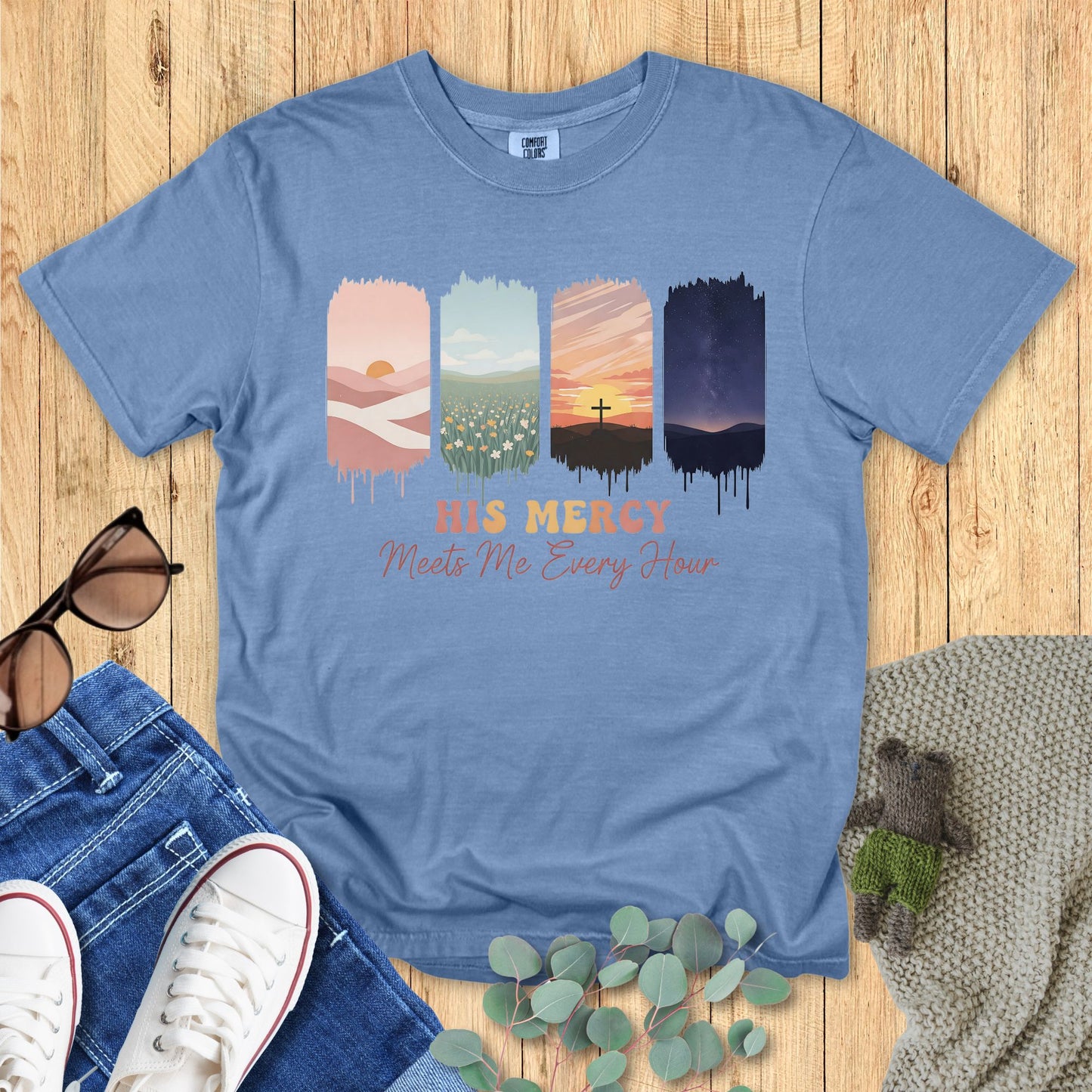 Comfort Colors Christian t-shirt in blue jean with Christian design “His Mercy Meets Me Every Hour.” Artwork shows four scenic panels—soft desert sunrise, floral field, cross at sunset, and starry night—symbolizing God’s mercy throughout the day. Flat lay mockup with denim, sneakers, and eucalyptus leave