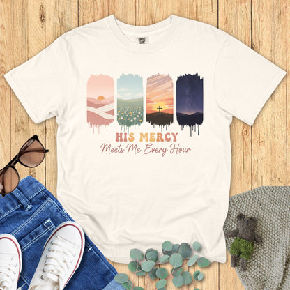 Comfort Colors Christian t-shirt in ivory with faith graphic “His Mercy Meets Me Every Hour.” Features four vertical painted panels depicting desert sunrise, blooming flowers, cross at sunset, and night sky. Christian apparel flat lay with casual outfit styling including jeans, sneakers, sunglasses, and greenery.