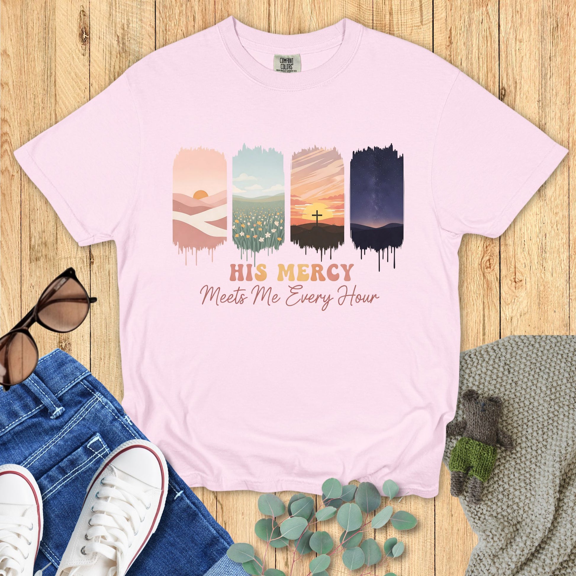 Comfort Colors Christian t-shirt in orchid purple with inspirational design “His Mercy Meets Me Every Hour.” Graphic features four vertical panels: sunrise desert hills, tulip field, sunset cross on a hill, and night sky with stars. Faith-based apparel styled flat lay with jeans, sneakers, sunglasses, and cozy knit