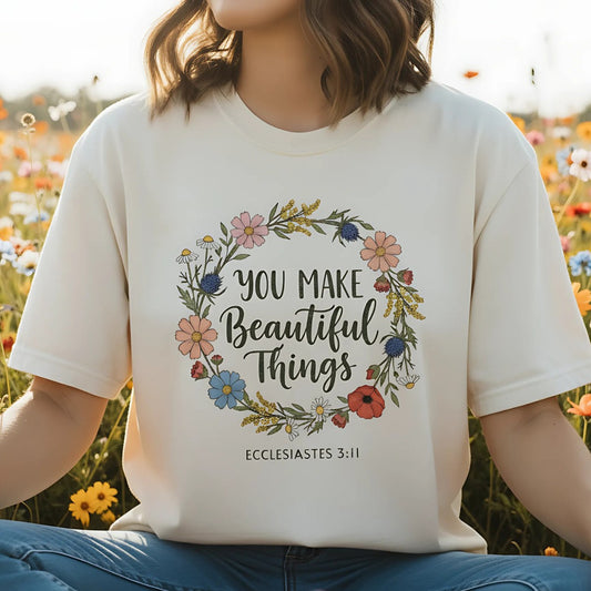 Woman sitting in a flower field wearing an ivory Comfort Colors Christian t-shirt with the Bible verse Ecclesiastes 3:11. The shirt features a colorful floral wreath design surrounding the phrase “You Make Beautiful Things.”