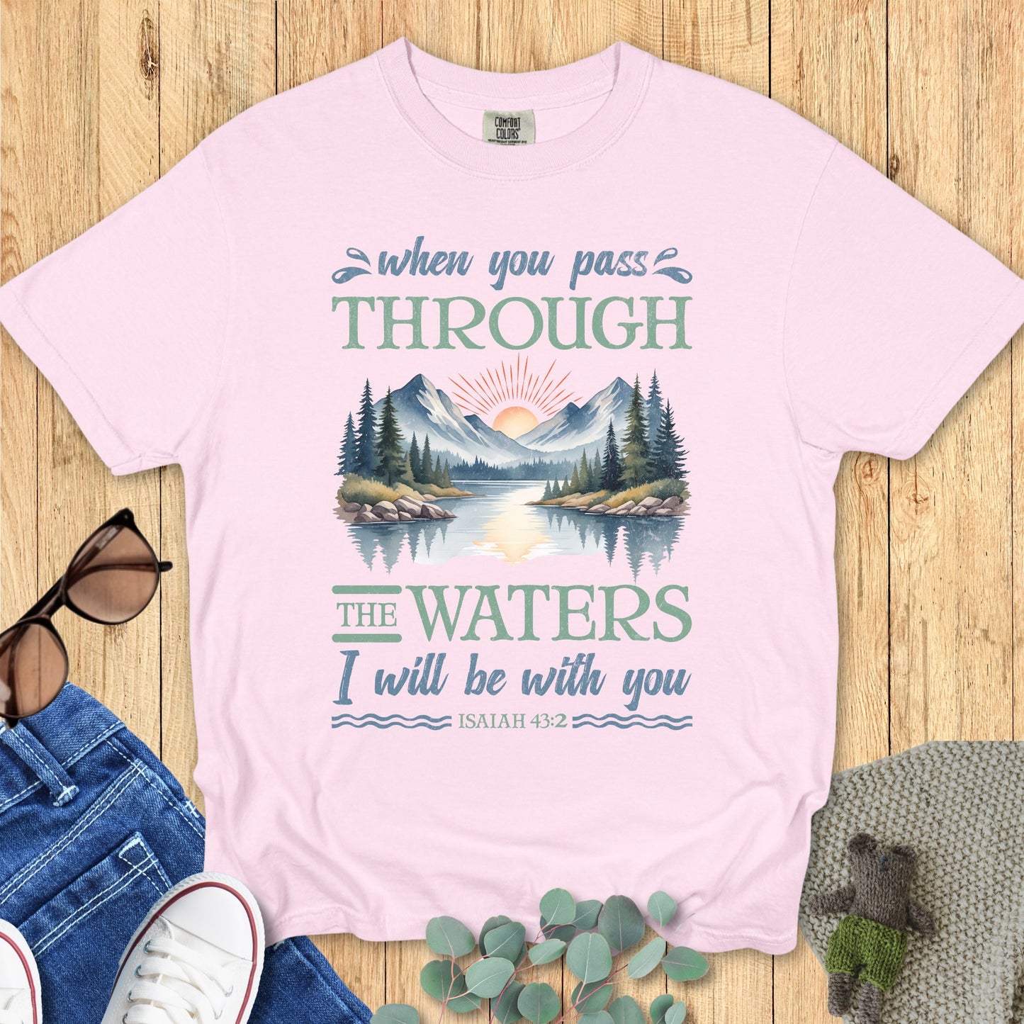 Comfort Colors Christian T-shirt in blossom pink featuring Isaiah 43:2 scripture. Illustrated graphic of mountains and water with sunrise, paired with the words “When you pass through the waters I will be with you.” Displayed flat with casual jeans, shoes, and accessories.