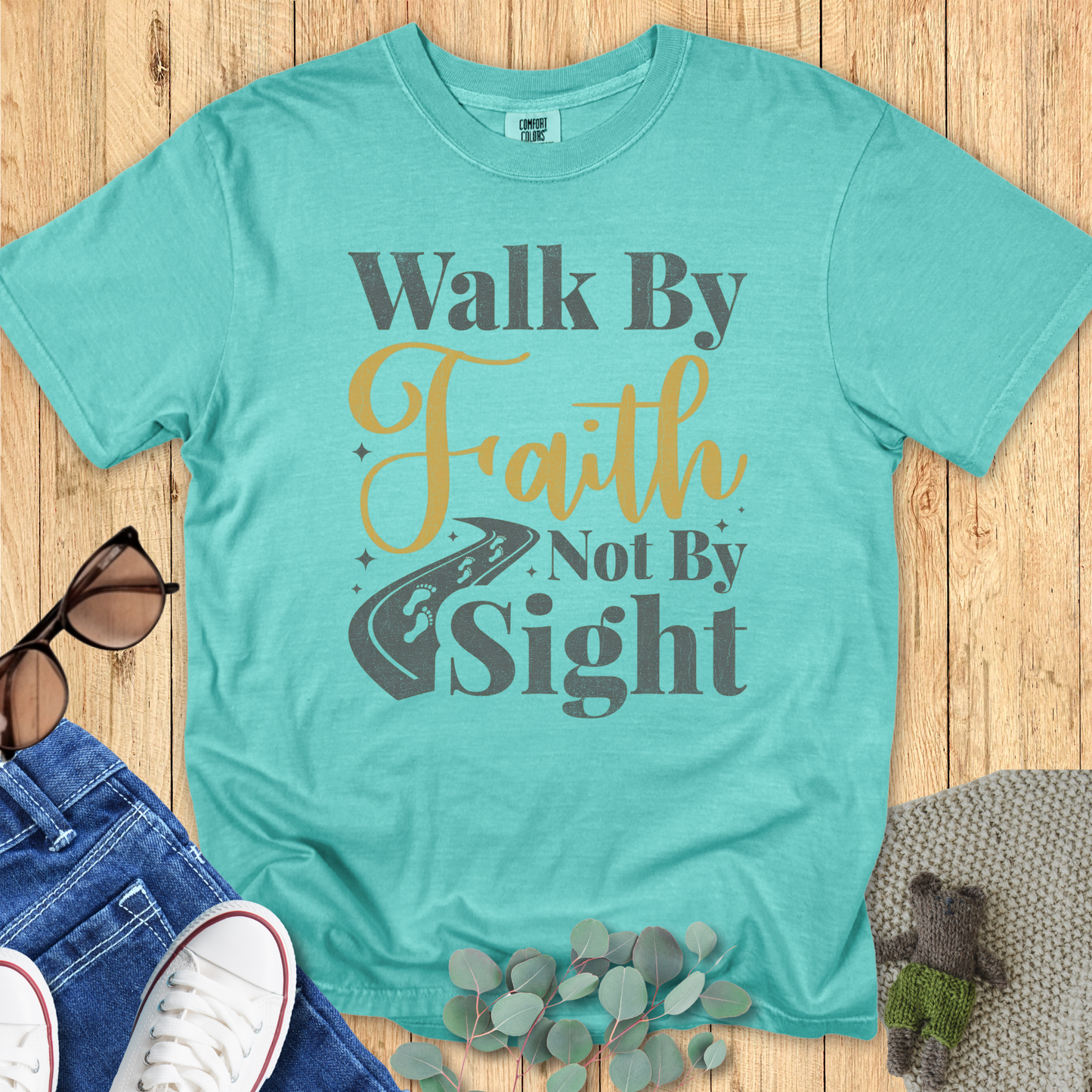 Seafoam Comfort Colors Christian t-shirt laid flat with the inspirational phrase 'Walk By Faith Not By Sight' in gold and gray print, road and footprints illustration, styled alongside denim, sneakers, and eucalyptus accents