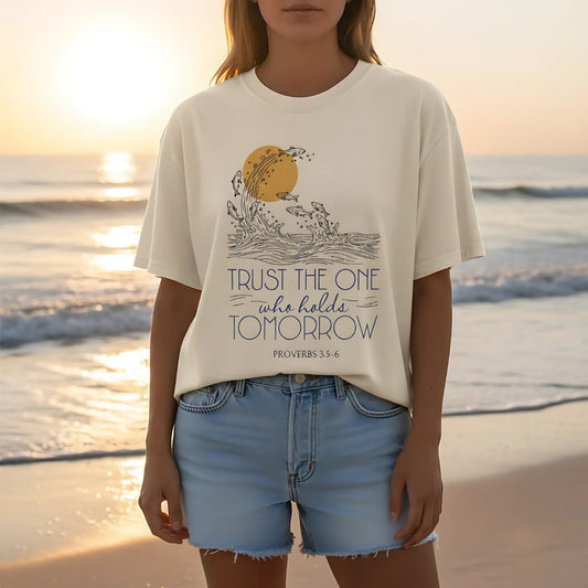 Woman wearing a natural ivory Comfort Colors Christian t-shirt on the beach at sunset. The Proverbs 3:5-6 design shows waves, fish, and a golden sun, paired with the words “Trust the One Who Holds Tomorrow”. Styled with denim shorts, creating a relaxed faith-inspired look