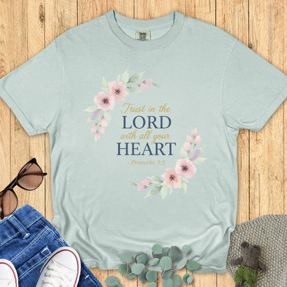 Flat lay of a soft bay green Comfort Colors Christian t-shirt with pastel pink floral graphics framing the Bible verse “Trust in the Lord with all your heart – Proverbs 3:5,” styled with jeans and sneakers