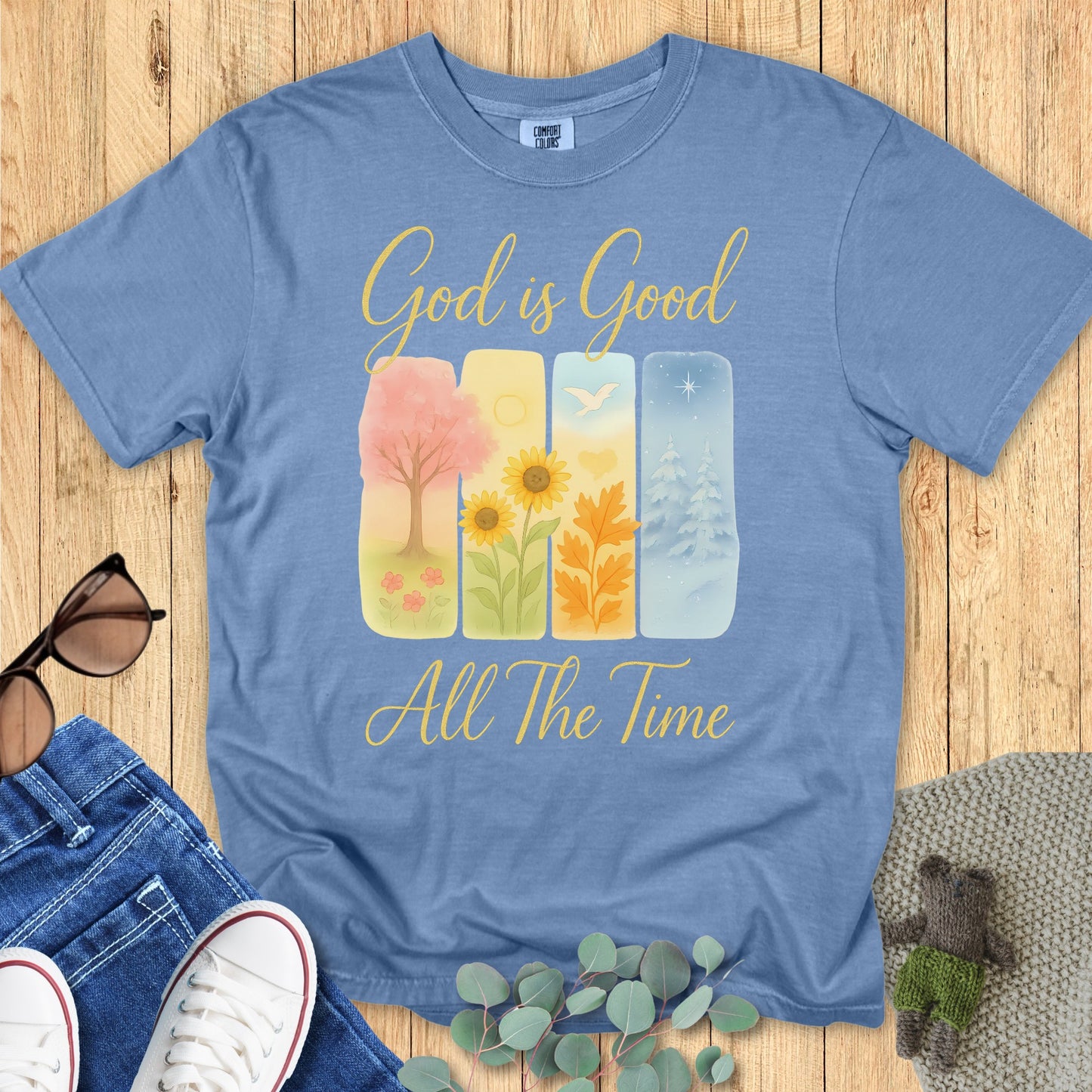 Comfort Colors 1717 Christian women t-shirt in blue jean, decorated with the Christian message "God is Good All The Time." The design includes four illustrated panels representing the seasons, symbolizing God's goodness year-round. Displayed in a casual flat lay with denim and accessories