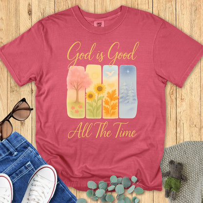 Comfort Colors 1717 Christian women t-shirt in crimson, printed with the phrase "God is Good All The Time" above seasonal nature artwork—spring tree, summer flowers, autumn leaves, and winter snow. Presented in a styled flat lay with casual lifestyle accessories.