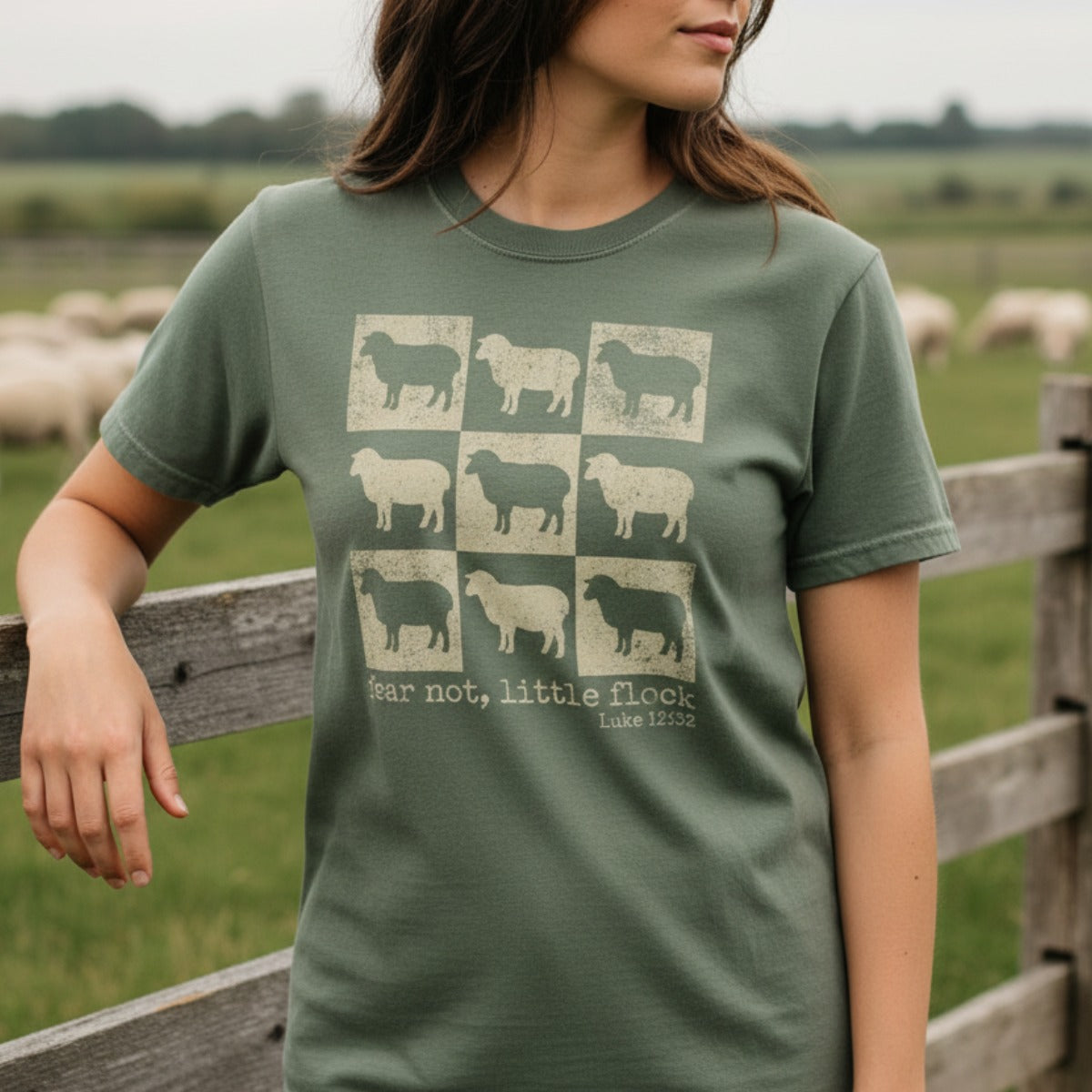 Christian women wearing a Comfort Colors 1717 moss green t-shirt featuring sheep grid design with scripture "fear not, little flock – Luke 12:32," standing outdoors by a fence with sheep in the background.