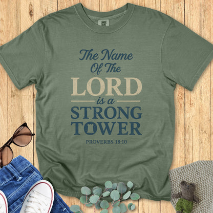 Flatlay of a Comfort Colors 1717 moss green “The Name of the Lord is a Strong Tower – Proverbs 18:10” t-shirt styled with jeans and white sneakers. A soft vintage piece from women’s scripture tees, perfect as a Christian mom gift idea.