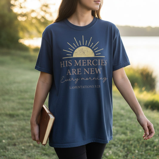 Woman holding a Bible and wearing a Comfort Colors 1717 midnight blue “His Mercies Are New Every Morning – Lamentations 3:23” t-shirt near sunrise. A peaceful Proverbs-inspired design for Christian mom gift ideas.