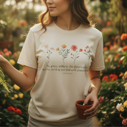 Woman tending plants in a greenhouse wearing a Comfort Colors 1717 ivory “The Grass Withers, The Flower Fades – Isaiah 40:8” t-shirt. A peaceful and elegant women’s scripture tee, making a thoughtful Christian mom gift idea.