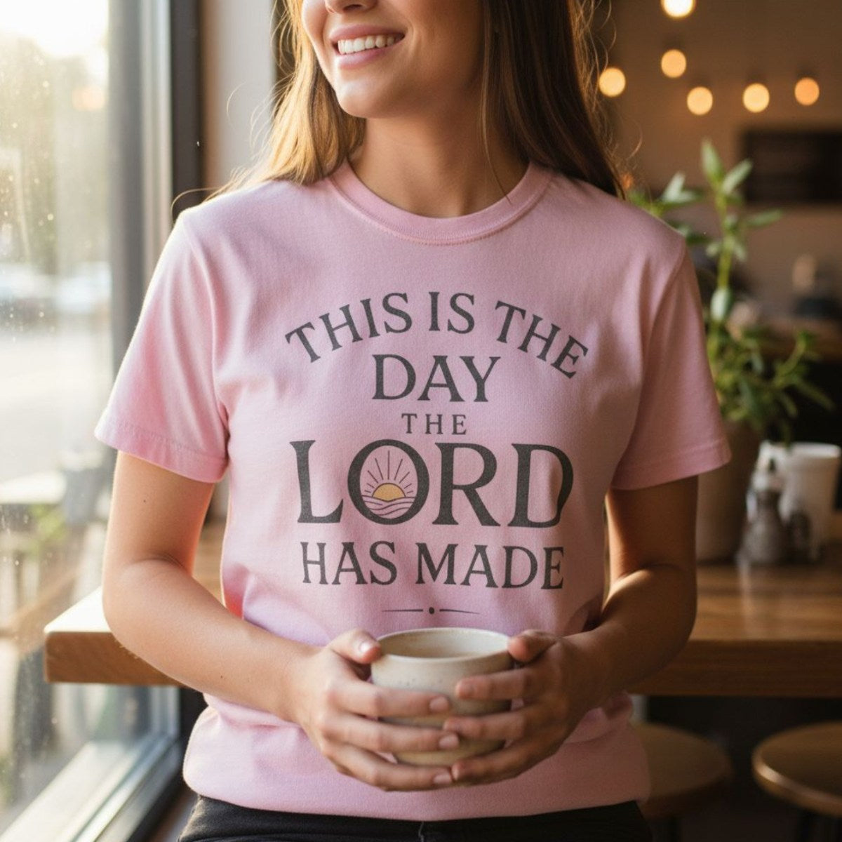 Woman sitting by a café window holding a coffee mug, wearing a Comfort Colors 1717 blossom pink “This Is The Day The Lord Has Made – Psalm 118:24” t-shirt. A joyful women’s scripture tee that makes a thoughtful Christian mom gift idea.