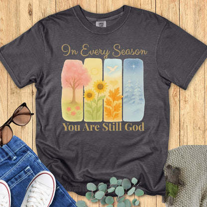 Flatlay of a Comfort Colors 1717 pepper “In Every Season You Are Still God” t-shirt featuring soft watercolor artwork of four seasons. A meaningful women’s scripture tee and faith-based apparel for women, perfect for everyday encouragement.
