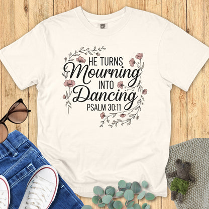 Flatlay of a Comfort Colors 1717 ivory “He Turns Mourning Into Dancing – Psalm 30:11” t-shirt decorated with soft blush flowers. A comforting faith-based apparel piece for women and meaningful Christian mom gift expressing hope and restoration.