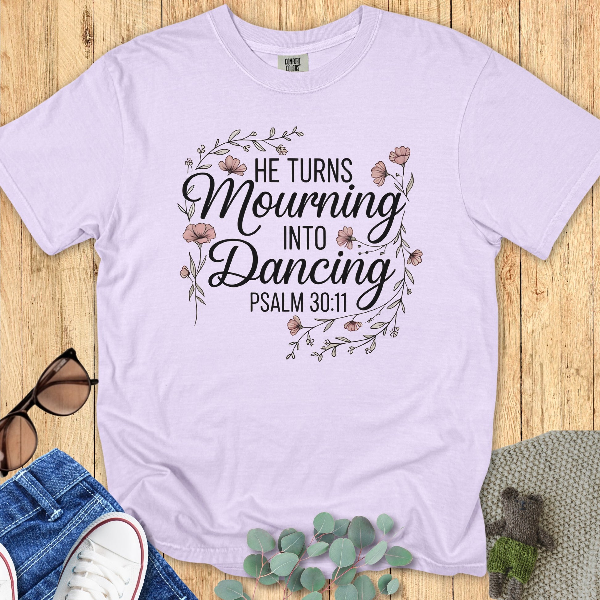 Flatlay of a Comfort Colors 1717 orchid “He Turns Mourning Into Dancing – Psalm 30:11” t-shirt featuring delicate pink floral accents. A soft and inspiring women’s scripture tee — uplifting faith-based apparel for women and a thoughtful Christian mom gift idea.