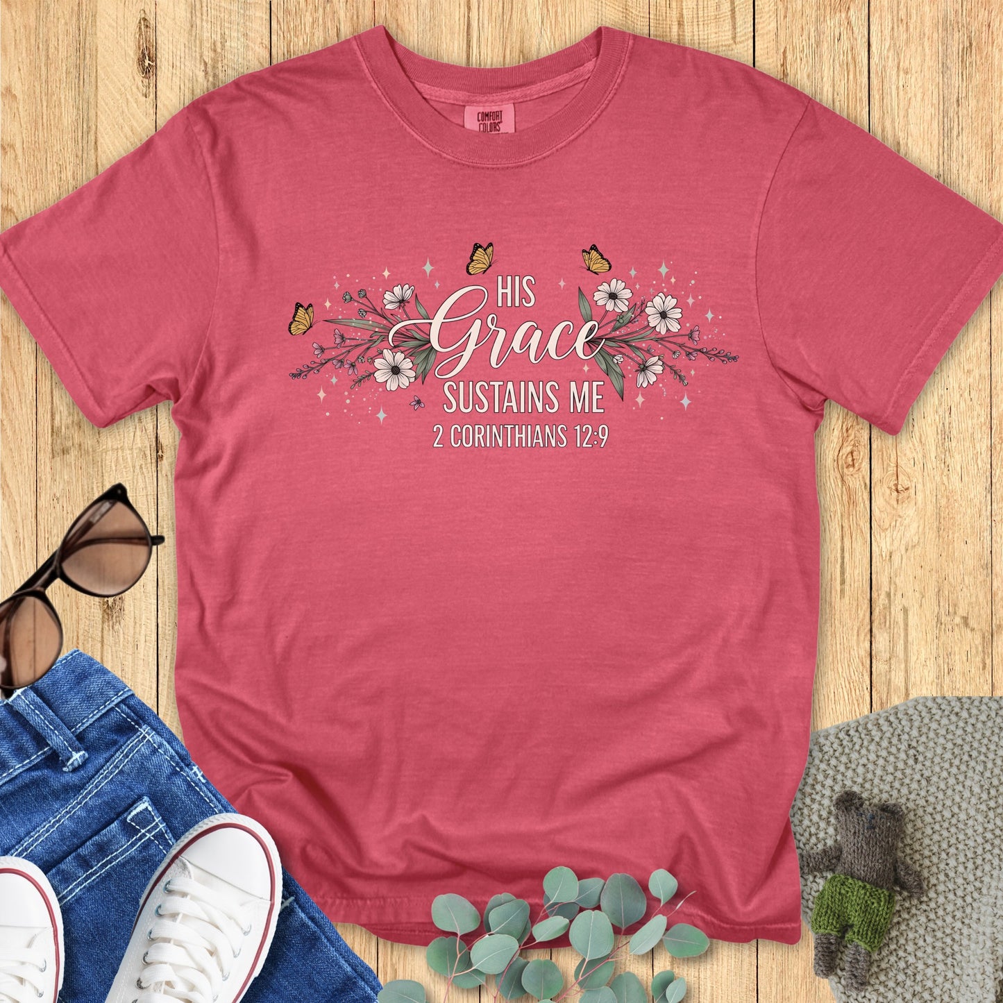 Flatlay of a Comfort Colors 1717 crimson “His Grace Sustains Me – 2 Corinthians 12:9” t-shirt decorated with elegant blossoms and butterflies. A meaningful piece of faith-based apparel for women — ideal Christian mom gift idea or daily encouragement tee.