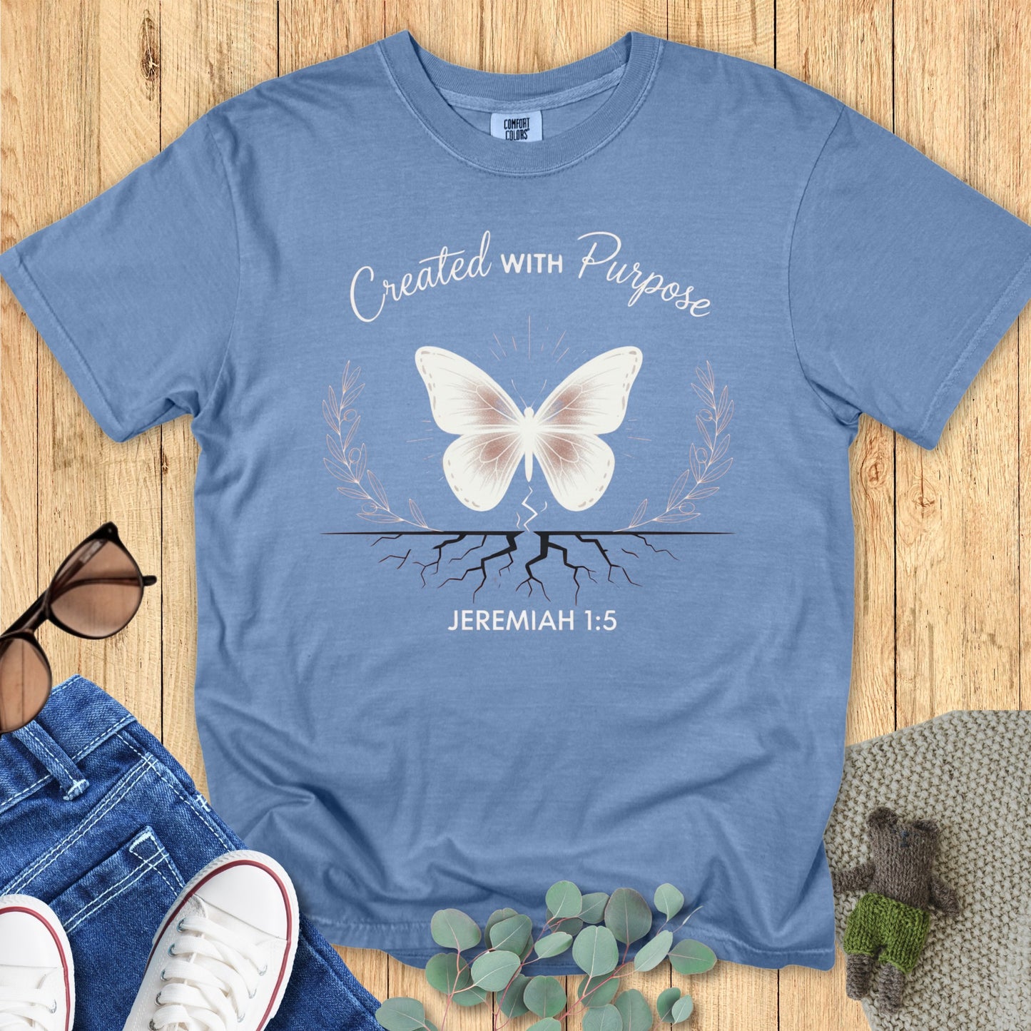 Flatlay of a Comfort Colors 1717 blue jean “Created With Purpose – Jeremiah 1:5” t-shirt featuring a butterfly above roots framed by laurel leaves. A soft, empowering piece of faith-based apparel for women — ideal Christian mom gift or daily reminder of purpose.
