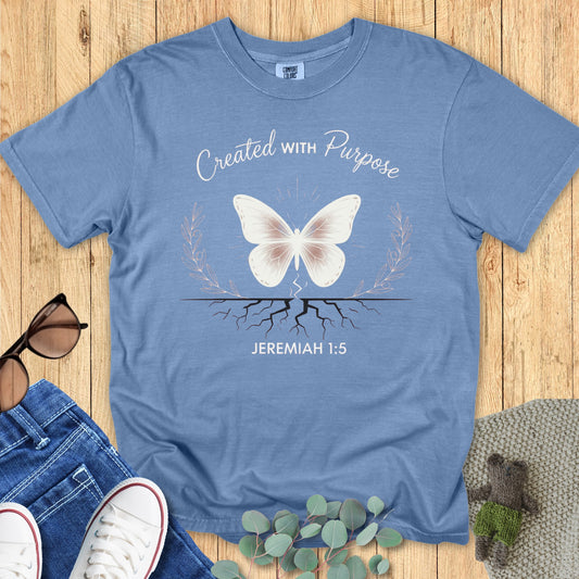Flatlay of a Comfort Colors 1717 blue jean “Created With Purpose – Jeremiah 1:5” t-shirt featuring a butterfly above roots framed by laurel leaves. A soft, empowering piece of faith-based apparel for women — ideal Christian mom gift or daily reminder of purpose.