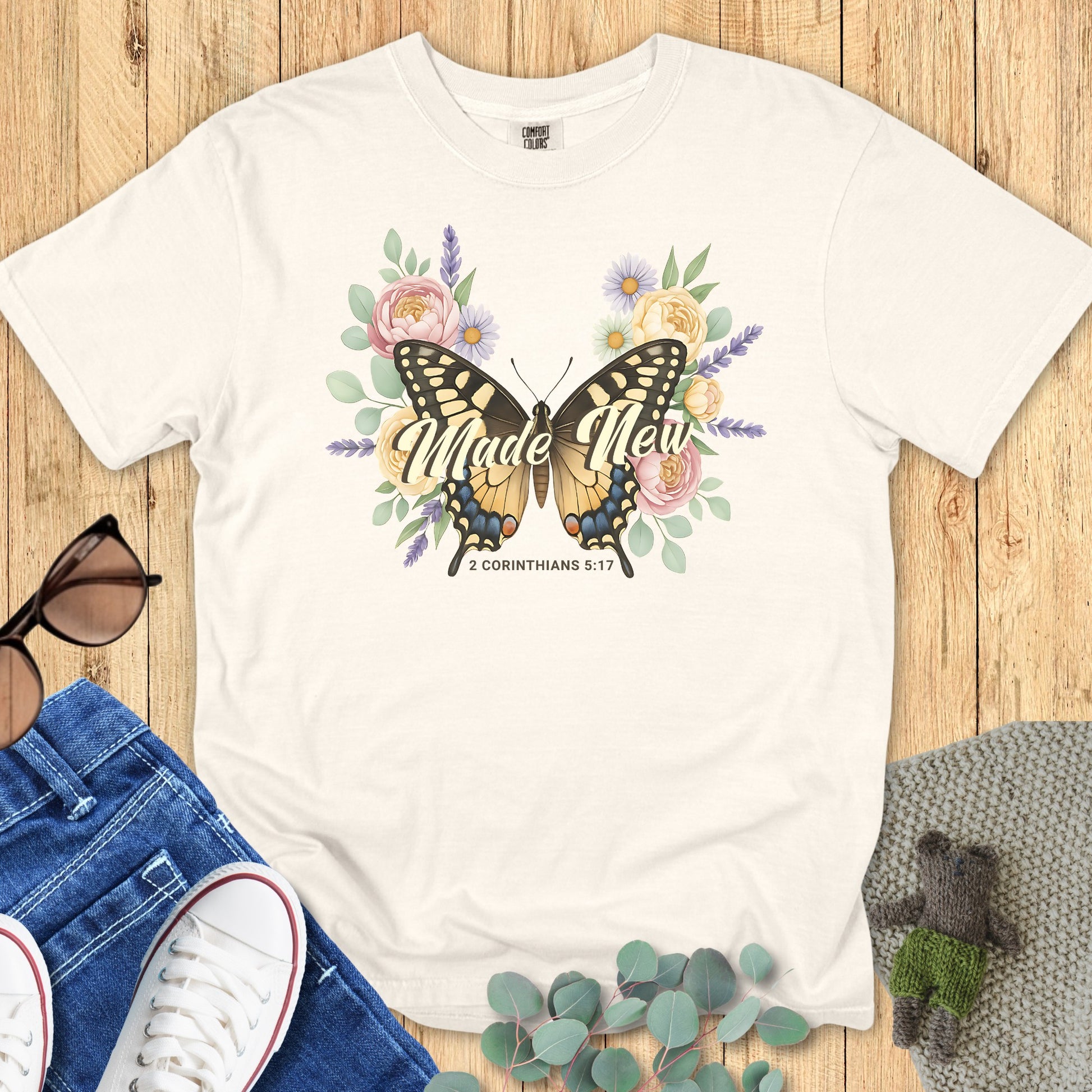 Flatlay of a Comfort Colors 1717 ivory “Made New – 2 Corinthians 5:17” t-shirt with vintage butterfly and floral illustration. A graceful and faith-filled women’s scripture tee — uplifting modern Christian shirt and meaningful gift for moms or believers.