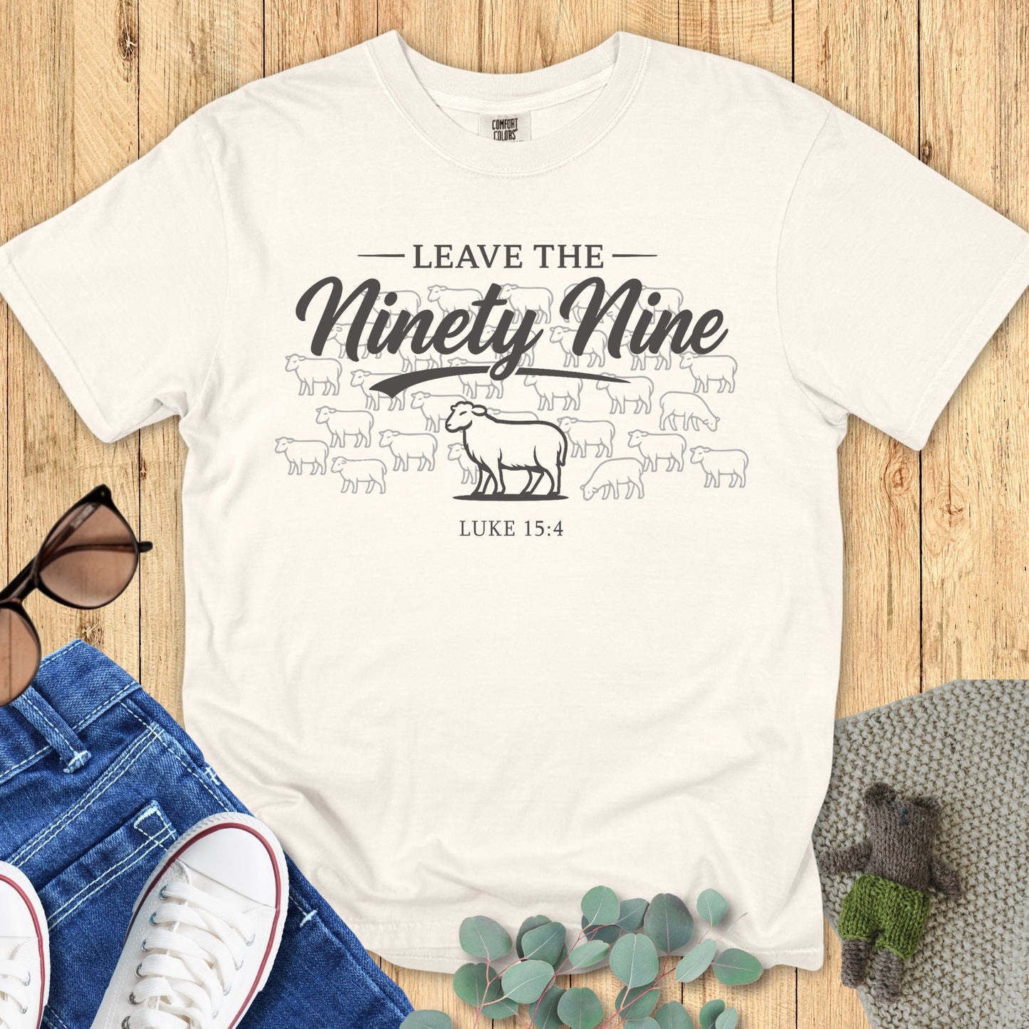 Flatlay of a Comfort Colors 1717 ivory “Leave the Ninety-Nine – Luke 15:4” t-shirt with minimalist sheep illustration. A simple yet powerful women’s scripture tee — elegant faith-based apparel for women and heartfelt Christian mom gift idea.
