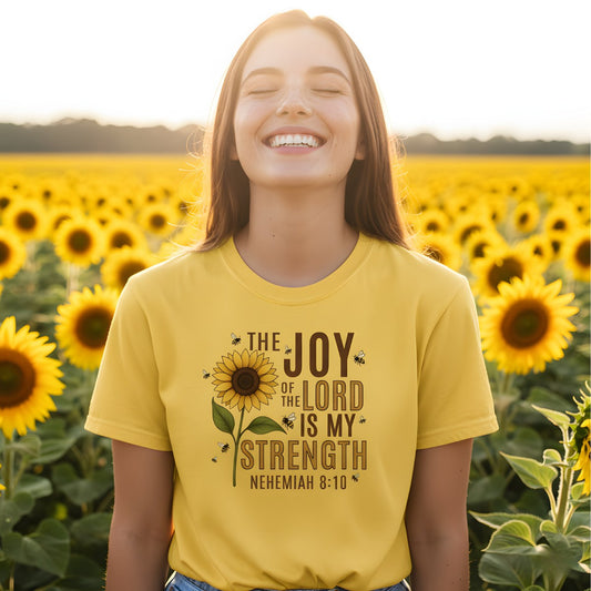 Smiling woman standing in a sunflower field wearing a Comfort Colors 1717 citrus “The Joy of the Lord Is My Strength – Nehemiah 8:10” t-shirt featuring a sunflower and bee design. A cheerful women’s scripture tee — modern Christian shirt and uplifting faith-based apparel for women, perfect Christian mom gift idea.