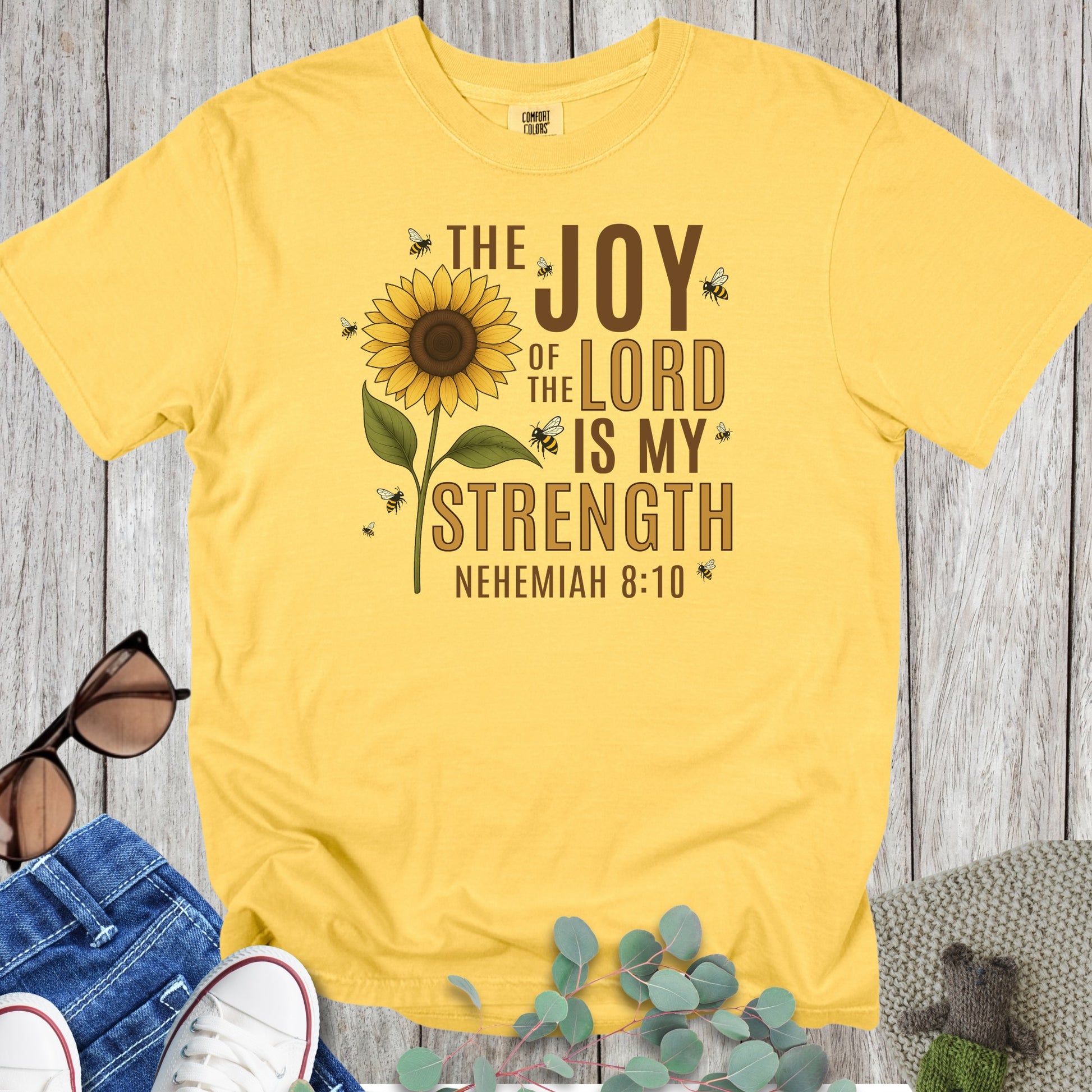 Flatlay of a Comfort Colors 1717 citrus “The Joy of the Lord Is My Strength – Nehemiah 8:10” t-shirt featuring a sunflower surrounded by bees. A radiant and joyful women’s scripture tee — bright faith-based apparel for women and thoughtful Christian mom gift.