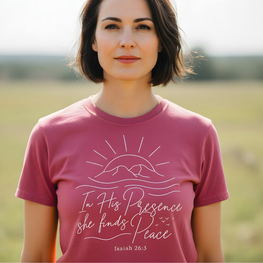 Woman standing outdoors wearing a Comfort Colors 1717 crimson “In His Presence She Finds Peace – Isaiah 26:3” t-shirt with elegant sunrise illustration. A serene women’s scripture tee — modern Christian shirt and faith-based apparel for women, perfect Christian mom gift idea.