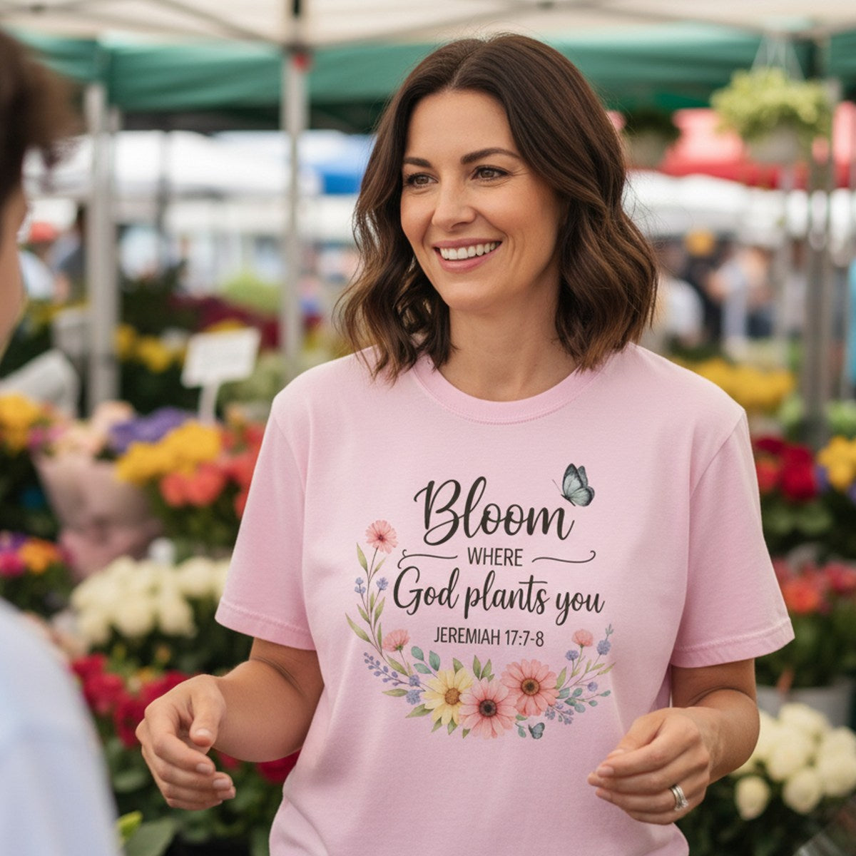 Woman smiling at a flower market wearing a Comfort Colors 1717 blossom “Bloom Where God Plants You – Jeremiah 17:7-8” t-shirt with floral and butterfly artwork. A joyful women’s scripture tee — stylish faith-based apparel for women and thoughtful Christian mom gift idea.