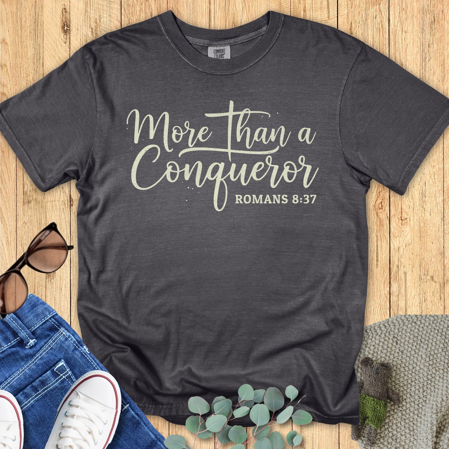Flatlay of a Comfort Colors 1717 pepper “More Than a Conqueror – Romans 8:37” t-shirt with soft cream lettering in elegant script. A bold women’s scripture tee — faith-based apparel for women and modern Christian shirt that makes a meaningful Christian mom gift idea.